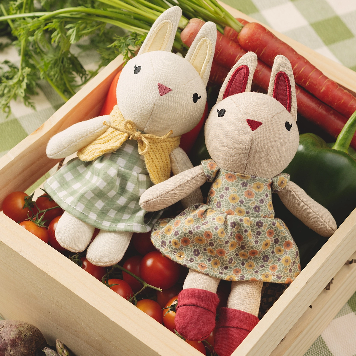 Two cream fabric rabbit plush toys dressed in patterned fabrics—one wearing a yellow bow and green gingham dress, the other in a floral print dress with burgundy socks—nestled in a wooden crate with fresh vegetables