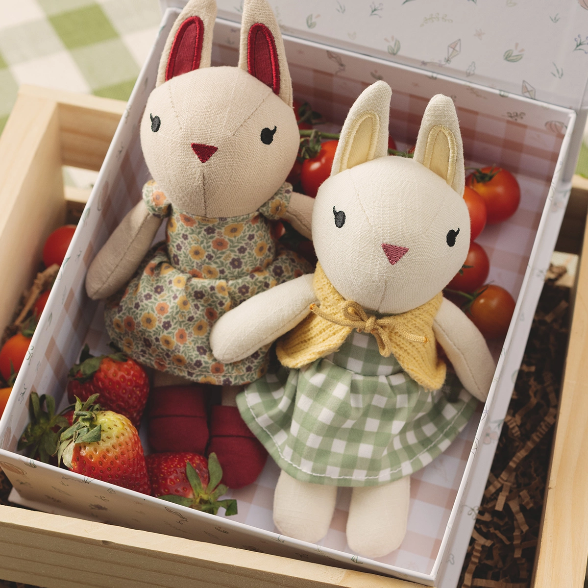 Two soft cream plush rabbits with embroidered faces and red inner ears, dressed in a floral print dress and yellow bow with gingham skirt, nestled in a display box with fresh strawberries and tomatoes