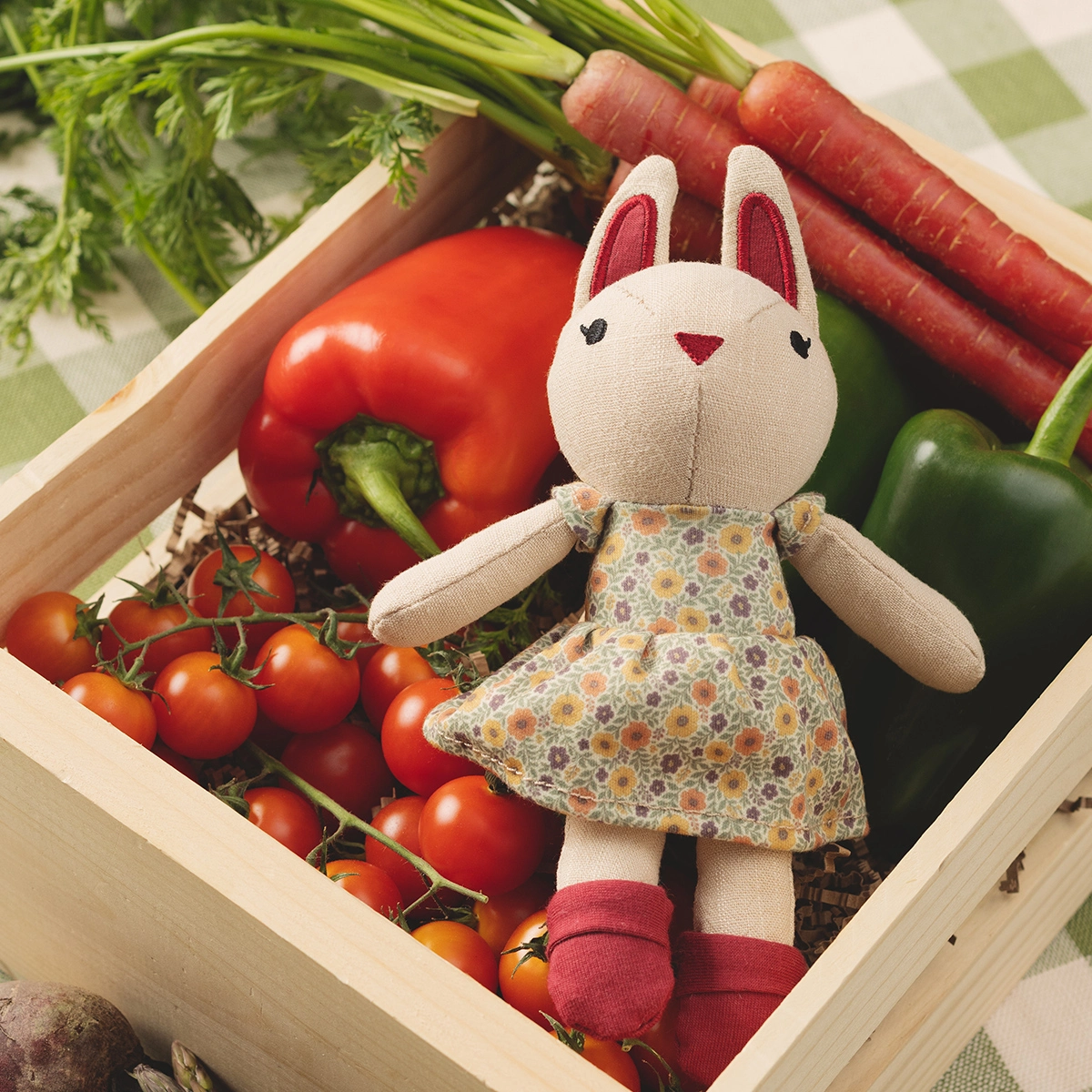 Cream fabric plush bunny with burgundy inner ears and nose, wearing a floral print dress in cream, orange and mauve tones, seated in a wooden crate surrounded by fresh vegetables including red tomatoes, carrots, bell pepper and leafy greens