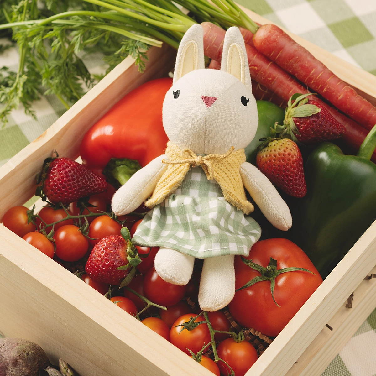 Cream plush bunny with embroidered black eyes and pink nose, wearing a yellow bow and green gingham dress, seated in a wooden crate surrounded by fresh tomatoes, strawberries, carrots and leafy greens