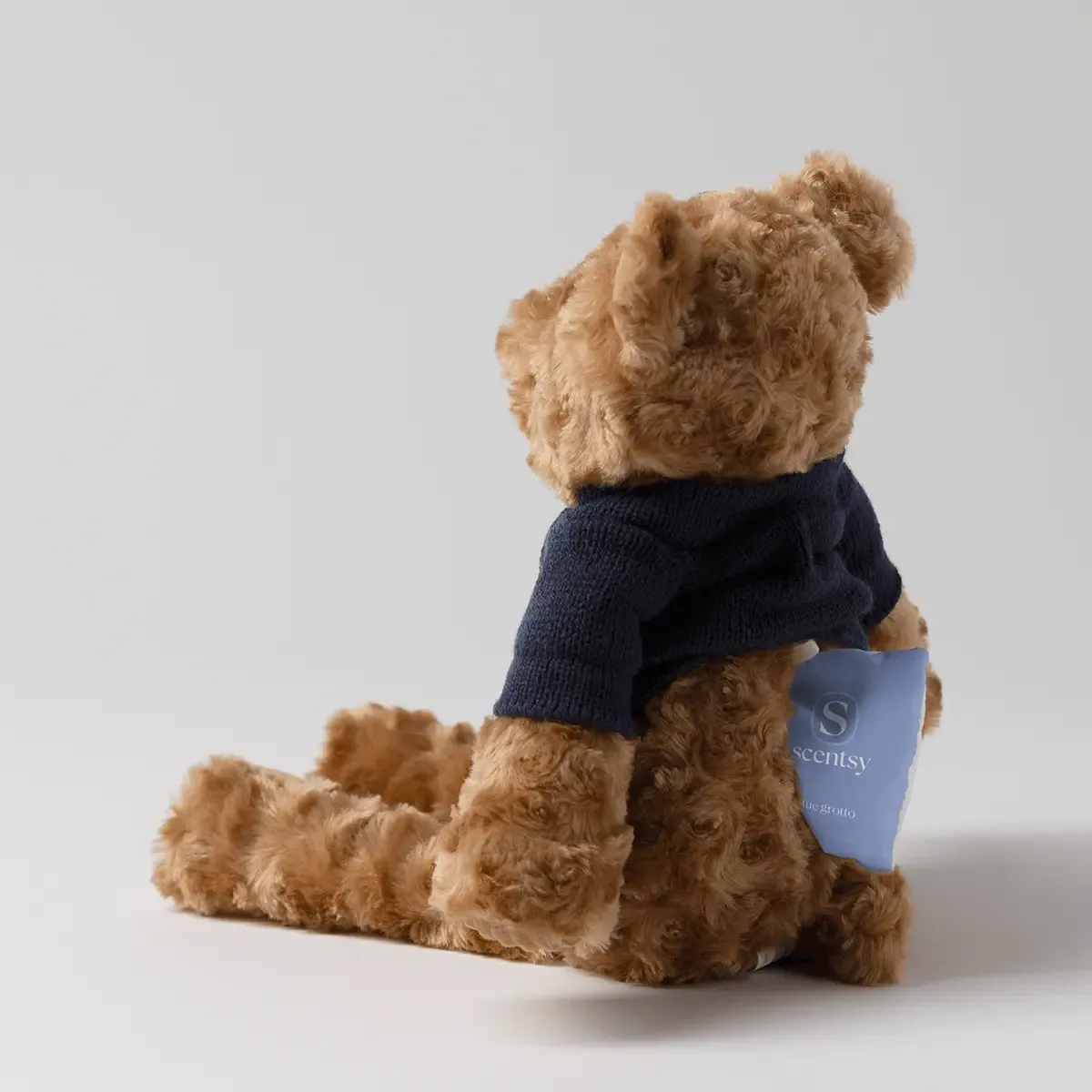 Tan plush teddy bear wearing a dark navy knit jumper, sitting with a blue Scentsy product tag attached to its arm