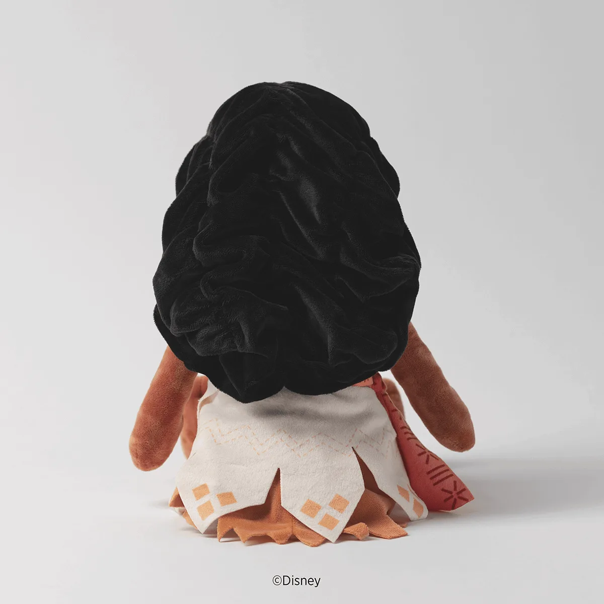 Soft plush toy with black curly hair, tan face and arms, and tan/cream skirt with orange diamond pattern details