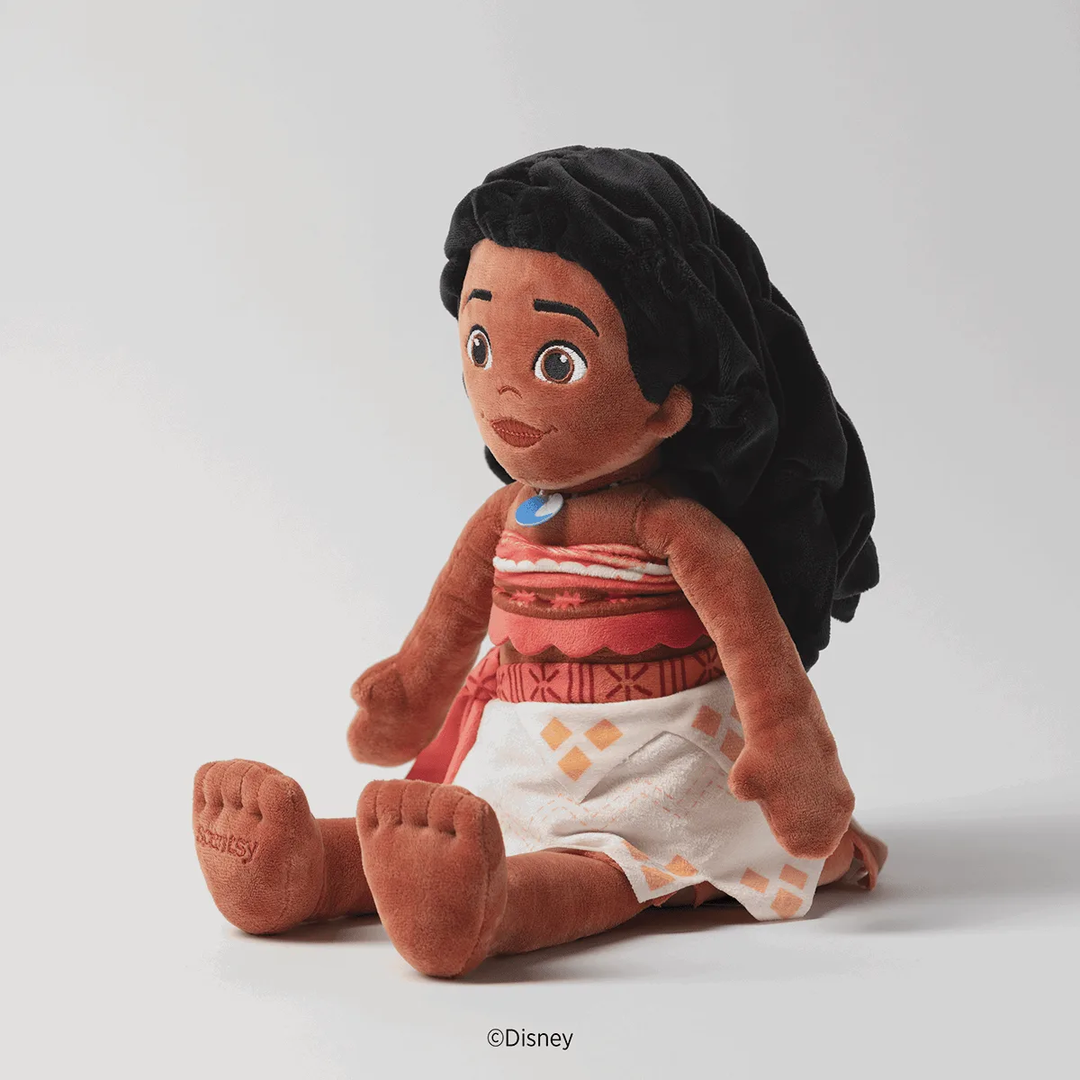Soft plush toy of Moana with long black hair, brown skin, wide eyes, and wearing her signature coral and white patterned dress with blue necklace pendant