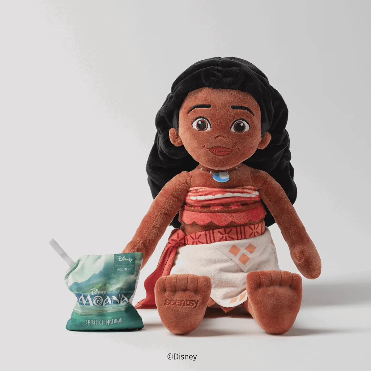 Soft plush Disney Moana Scentsy Buddy with black long hair, brown skin, and red and white traditional dress with blue necklace, sitting next to a green Moana-themed Scentsy Scent Pak pouch labelled Spirit of Motunui