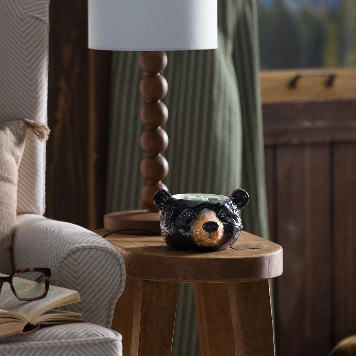 Black and tan bear head warmer with realistic facial features, sitting on a wooden side table next to a cream upholstered chair, the Blackwood Bear Scentsy Warmer