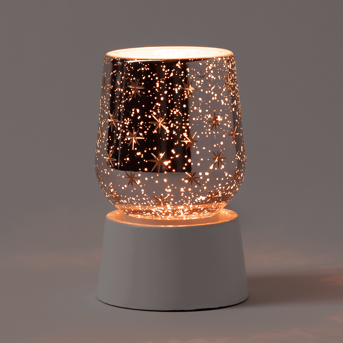 White ceramic tabletop base with rose gold perforated warmer featuring star cutouts and scattered light dots, illuminated from within