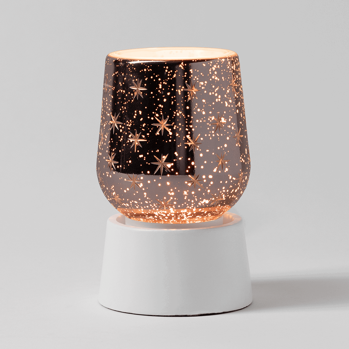Rose gold metal mini warmer with a dimensional starburst and sparkle cutout pattern that creates light effects, paired with a cylindrical white tabletop base, the Frosted Flurry Mini Scentsy Warmer with Tabletop Base