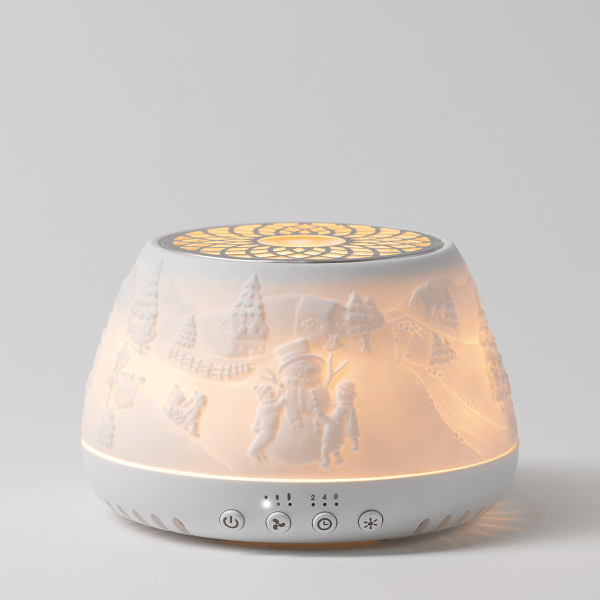 White ceramic diffuser with embossed winter scene featuring trees, figures and decorative elements, illuminated from within with warm golden light, four touch-control buttons on base, the Scentsy Air Quad - Icicle Lan