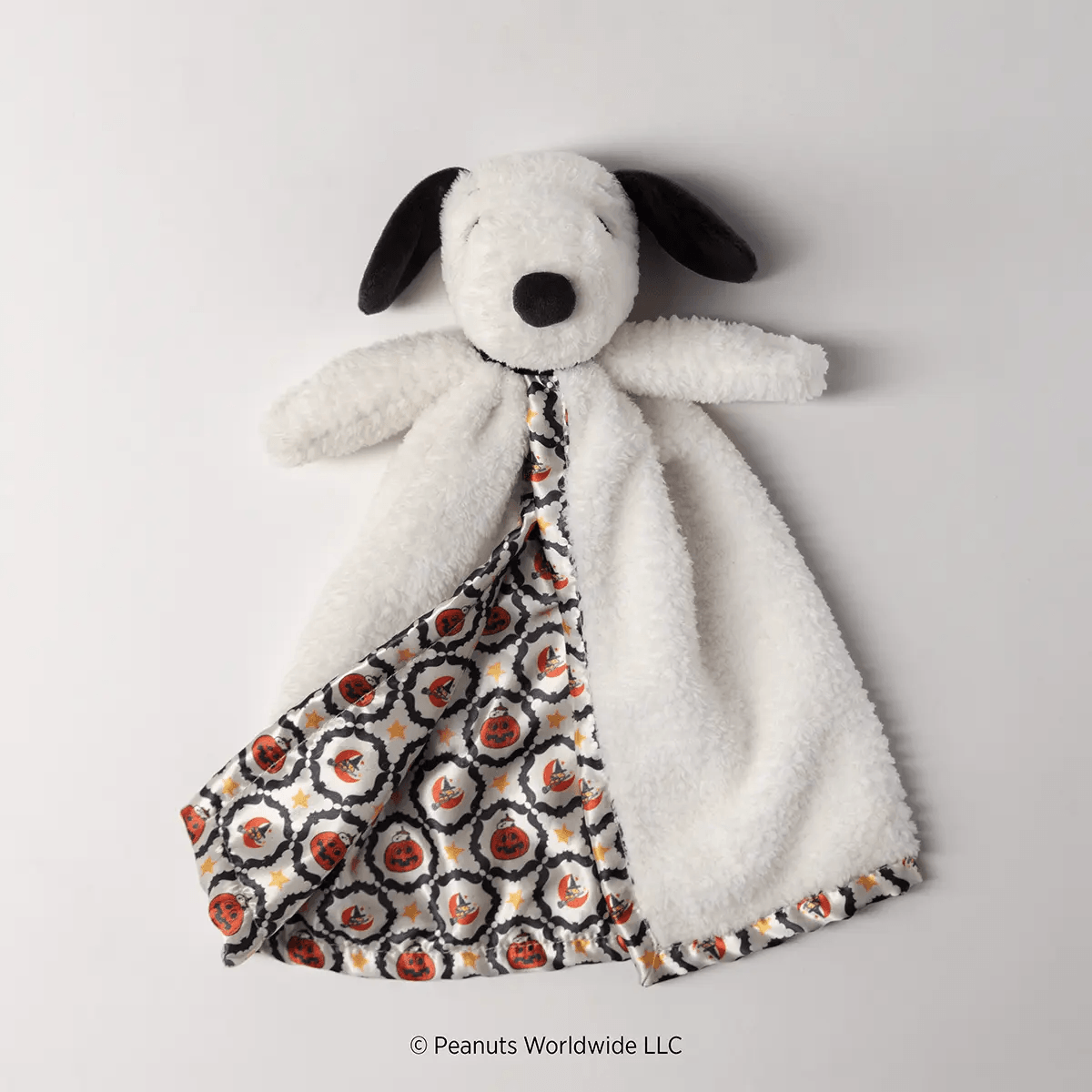 Soft white plush dog with black ears and nose wearing a patterned blanket with red pumpkins, yellow stars, and black swirls, Snoopy’s Happy Halloween Blankie Buddy