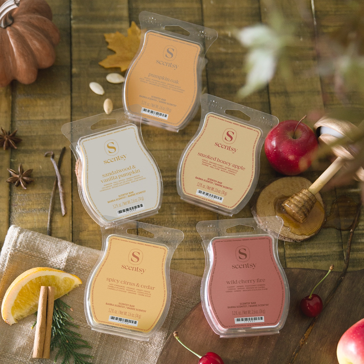 Five Scentsy Bars in autumn-themed scents arranged on a wooden surface with seasonal styling: pumpkin spice, sandalwood & vanilla pumpkin, smoked honey apple, spicy citrus & cedar, and wild cherry fire, each in clear clamshell packaging with the Scentsy logo