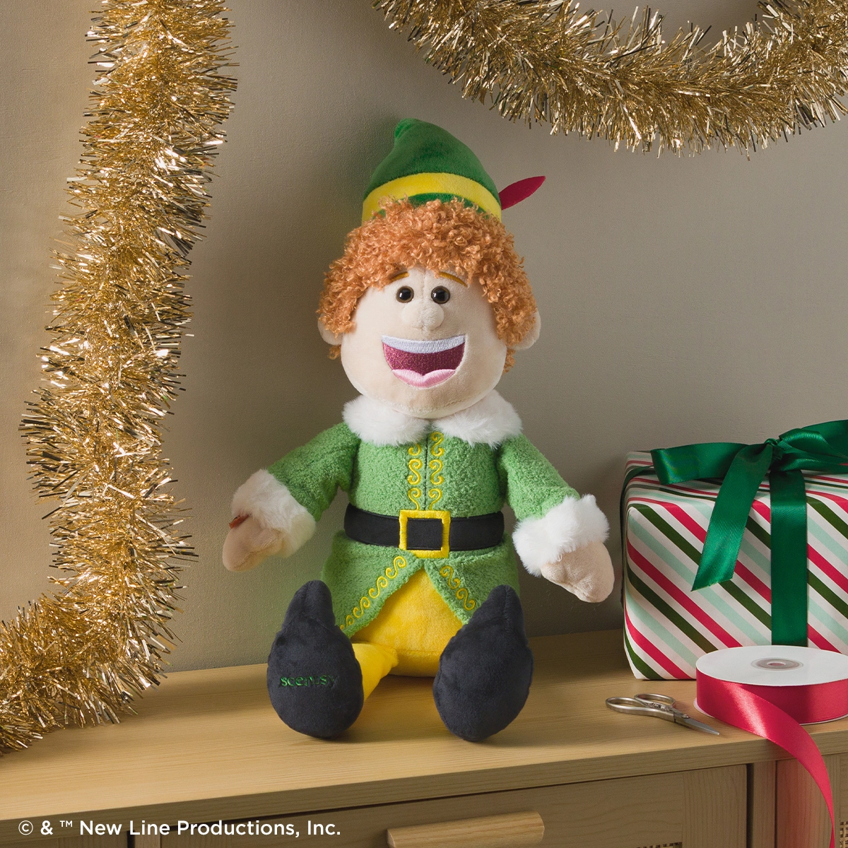 Plush elf character with curly ginger hair, wide grin, green jacket with yellow front panel and black belt, white-furred collar and cuffs, black shoes, and a green hat with yellow band and red feather, displayed on a shelf with gold tinsel and wrapped gift box