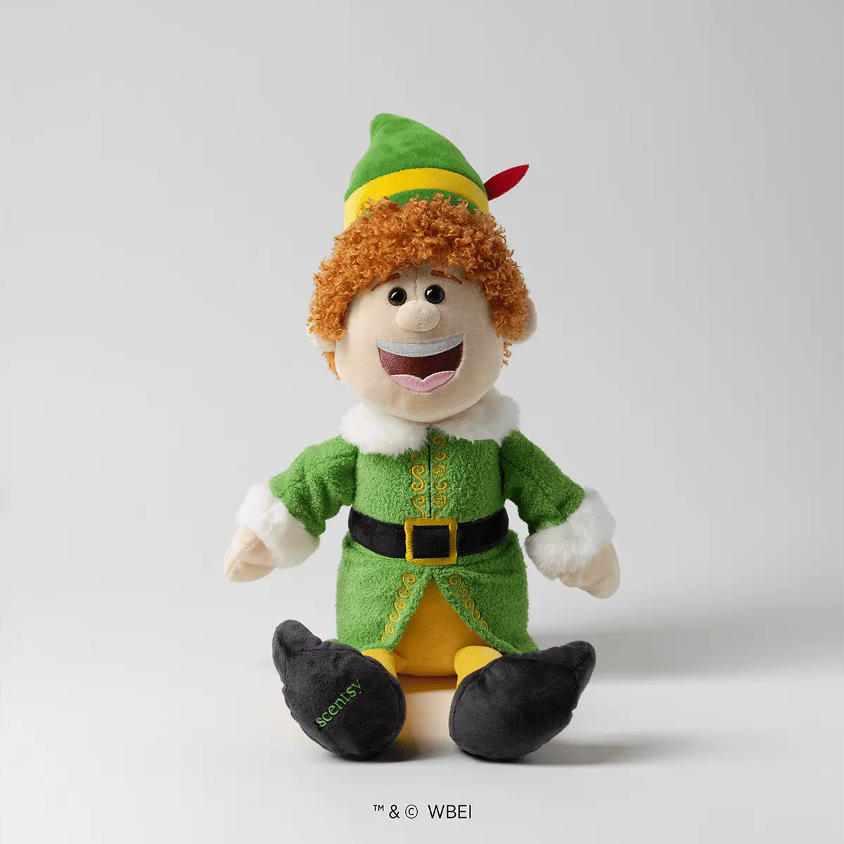 Smiling plush elf character with curly orange hair, green pointed hat with red feather, green tunic with gold embroidery and black belt, white collar and cuffs, and black shoes