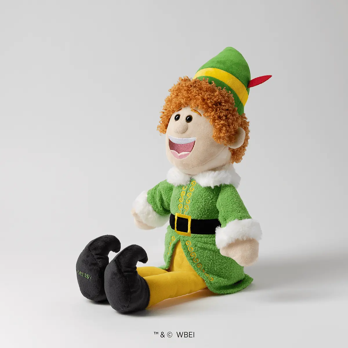 Smiling plush character with curly orange hair, wearing a green felt coat with yellow trim, white fur cuffs, a yellow belt with black buckle, green hat with red feather, and oversized black curled-toe shoes with yellow soles