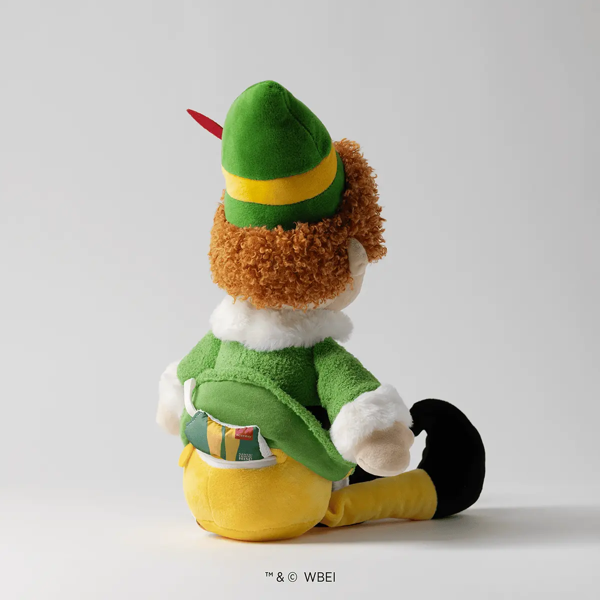 Plush toy elf with curly brown hair, green pointed hat with yellow band and red feather, green jacket with white collar and cuffs, yellow trousers, and black shoes, sitting in profile