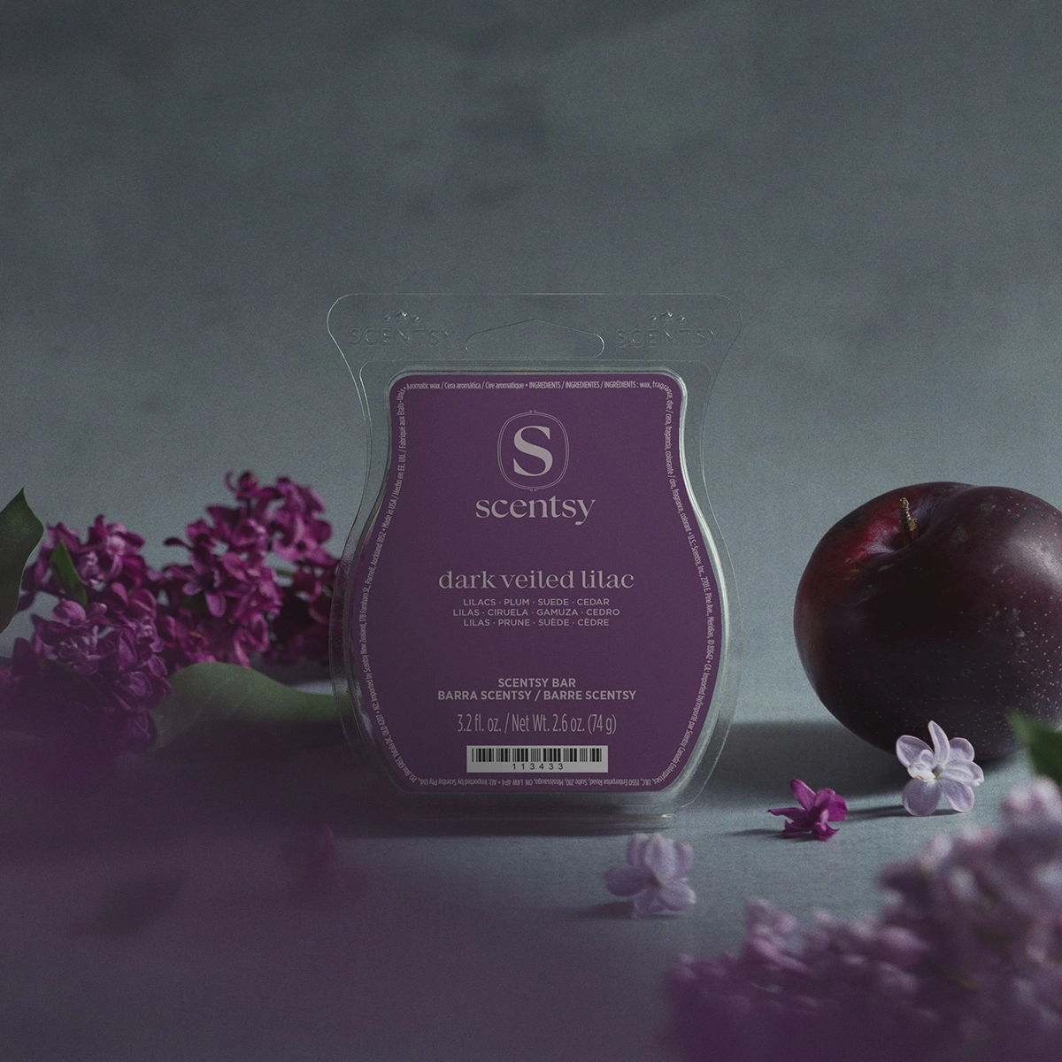 Clear clamshell packaging containing a purple Scentsy wax bar labelled Dark Veiled Lilac, surrounded by fresh purple lilac flowers, a dark plum, and white blossoms on a grey surface