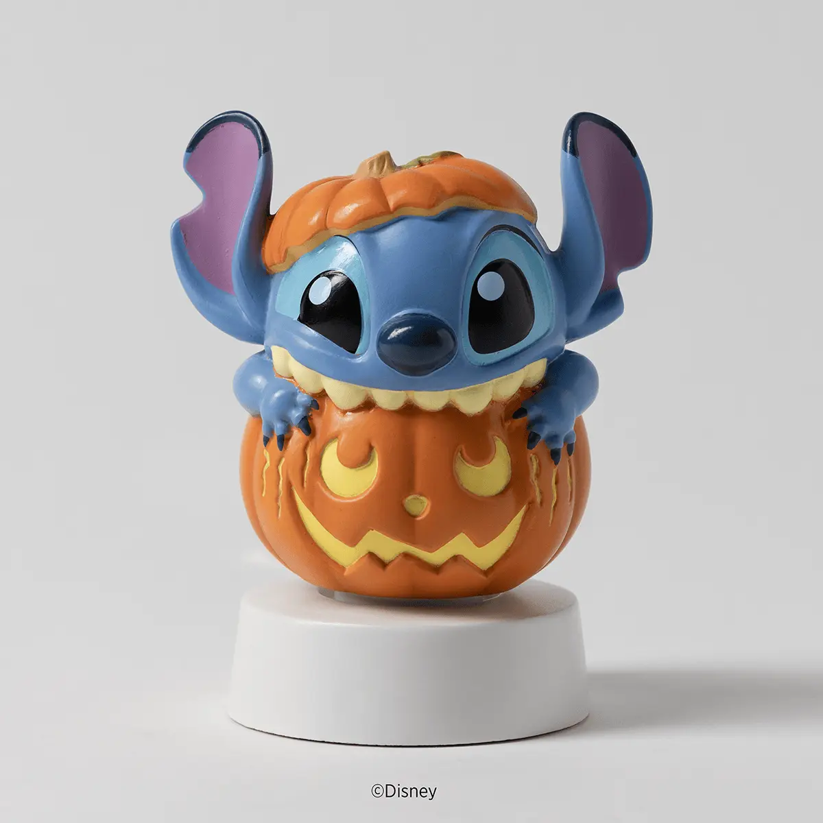 Blue alien character with large ears perched on top of an orange jack-o'-lantern with carved face details, mounted on a white circular base, the Disney Stitch: Pumpkin Trouble Mini Scentsy Warmer with Tabletop Base