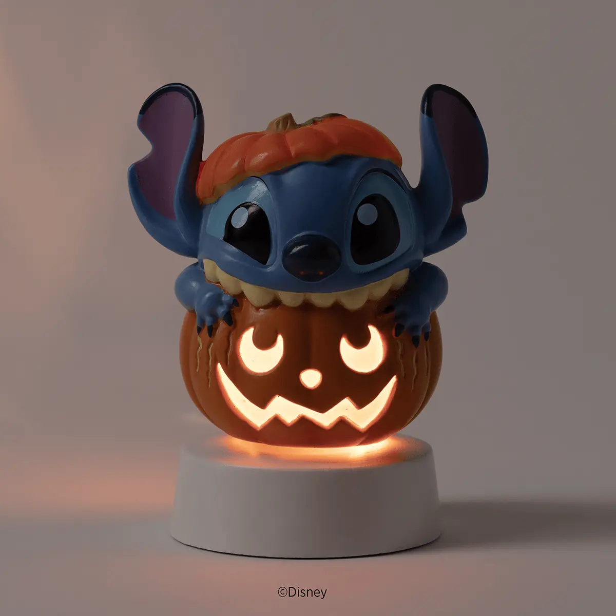 Blue alien character perched on a carved pumpkin with glowing jack-o'-lantern face, wearing an orange hat, on a white tabletop base