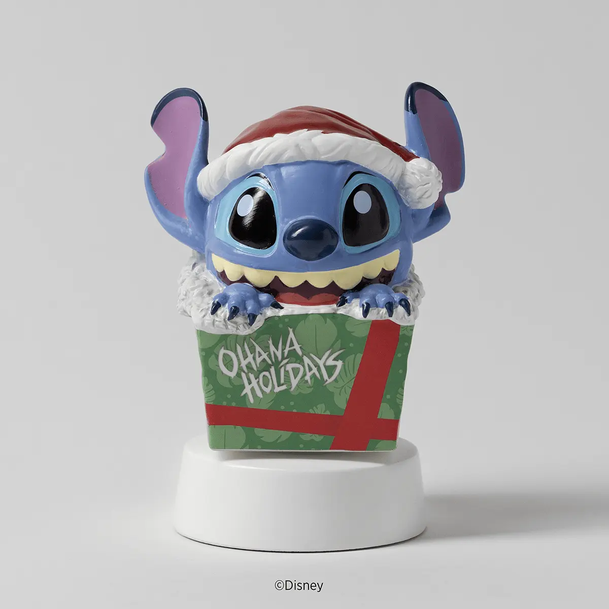 Blue character with large ears wearing a Santa hat and festive outfit, sitting in a green gift box with red ribbon and "Ohana Holidays" text on a white cylindrical base, Disney Stitch: Unleash the Holidays Mini Scentsy Warmer with Tabletop Base