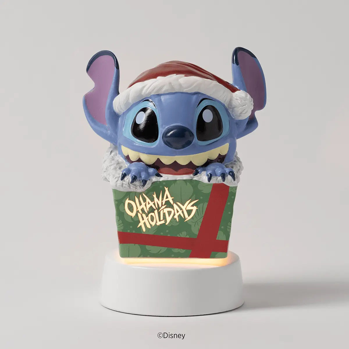 Blue Disney character with large ears wearing a Santa hat, seated in a green gift box with red ribbon and "Ohana Holidays" text, mounted on a white cylindrical base with interior lighting