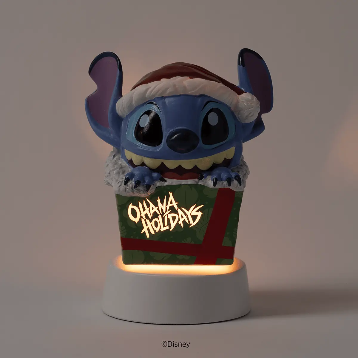 Blue alien character with large ears wearing a Santa hat and white fur trim, sitting in a green gift box with red ribbon and "Ohana Holidays" text, with illuminated base, the Disney Stitch: Unleash the Holidays Mini Scentsy Warmer