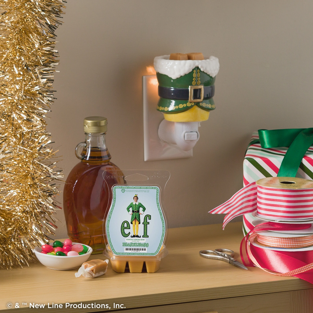 Elf: SANTA! I KNOW HIM! Scentsy Bar displayed on a wooden shelf surrounded by festive holiday décor including gold tinsel, ribbon spools, and seasonal ornaments