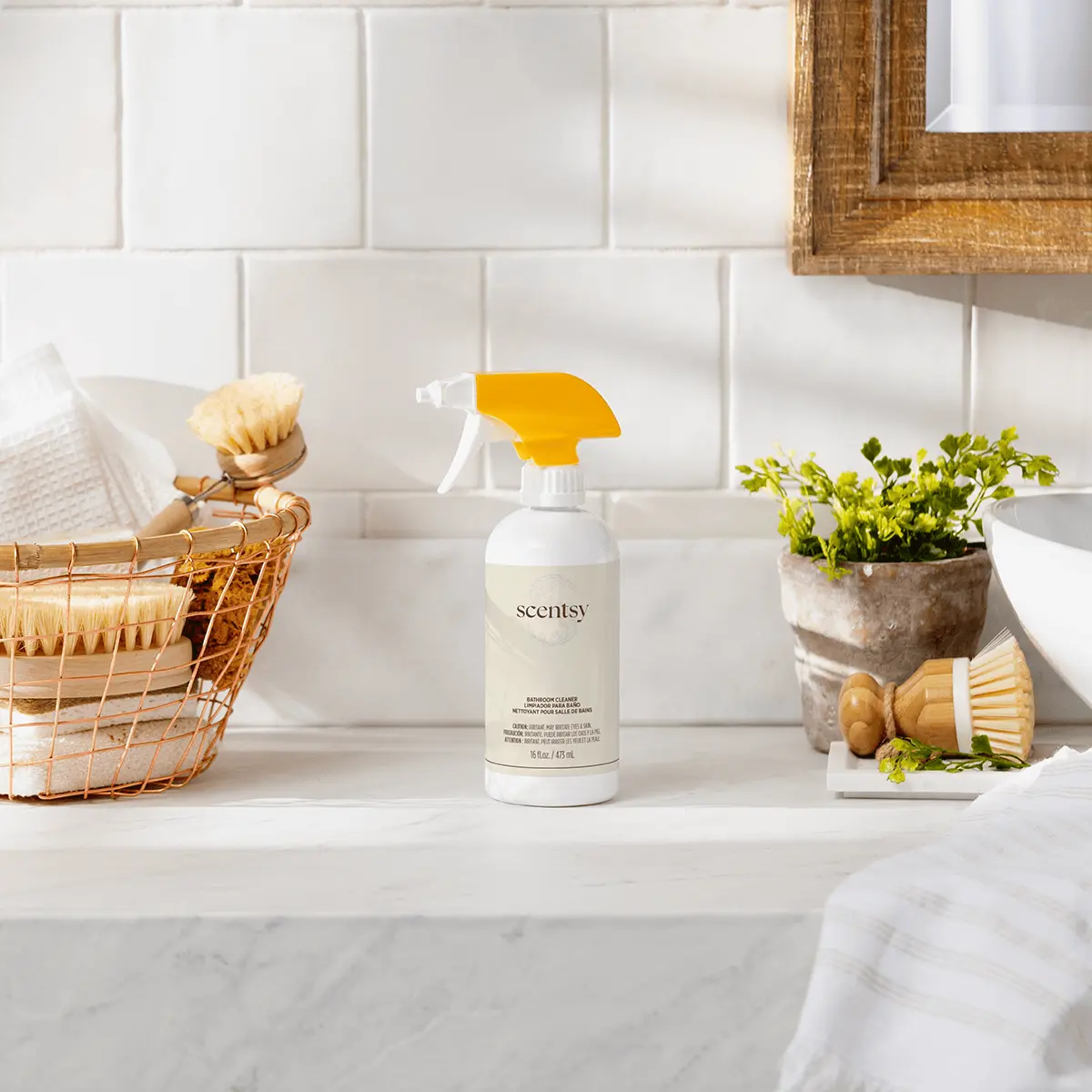 White spray bottle with yellow trigger and "Scentsy" label, the Frosted Eucalyptus Bathroom Cleaner, displayed on a marble countertop alongside cleaning brushes and potted herbs
