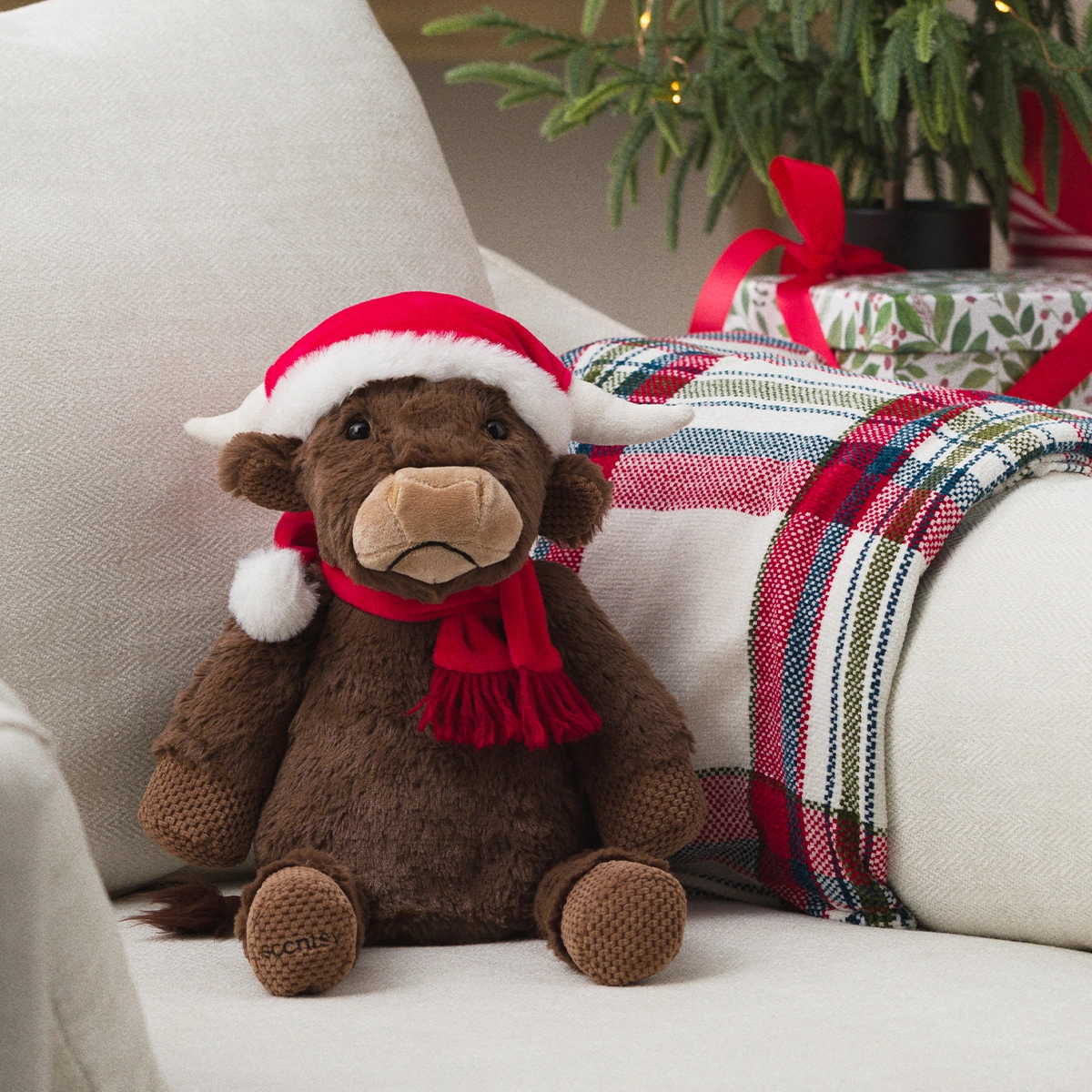 Brown plush bear wearing a red and white Santa hat and matching red scarf, seated on a cream cushion with festive tartan fabric and greenery in the background