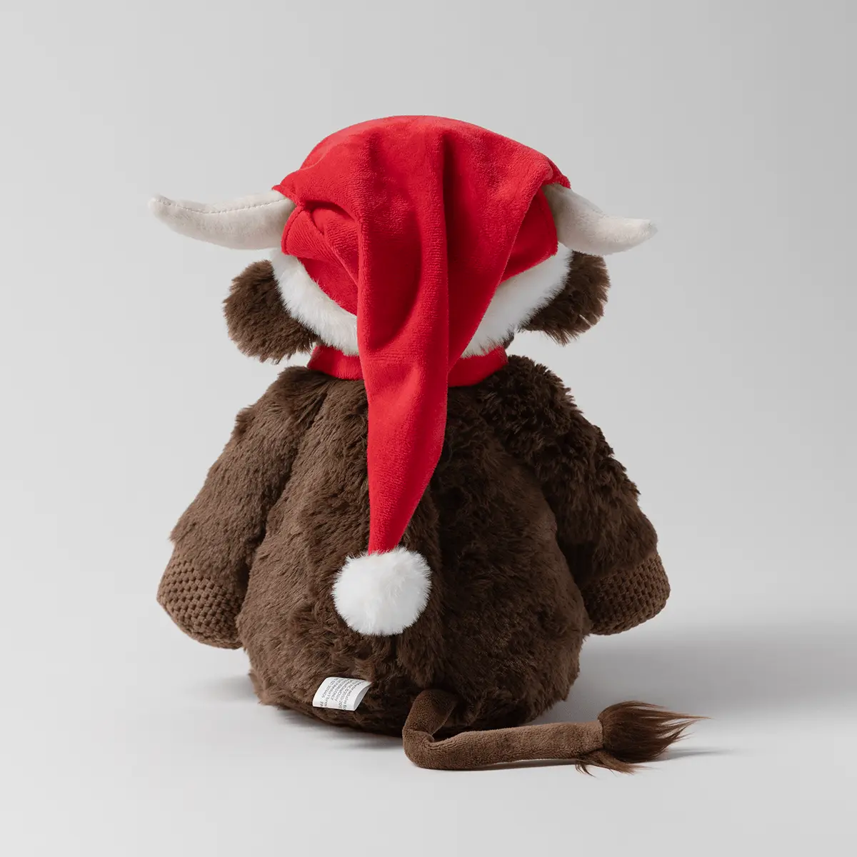 Plush cow character wearing a red Santa hat with white trim, cream horns, and a white pom-pom on its brown body, viewed from behind