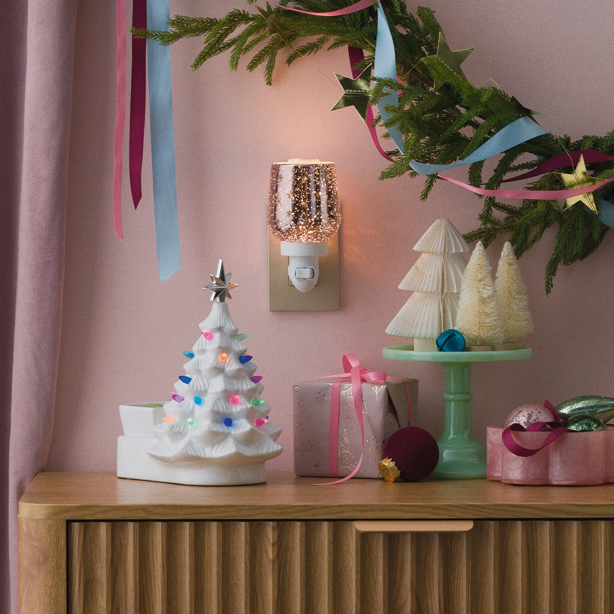 White ceramic Christmas tree warmer with colourful ornament details, displayed on a wooden dresser among festive decorations and greenery, the Merry Little Christmas - White Scentsy Warmer