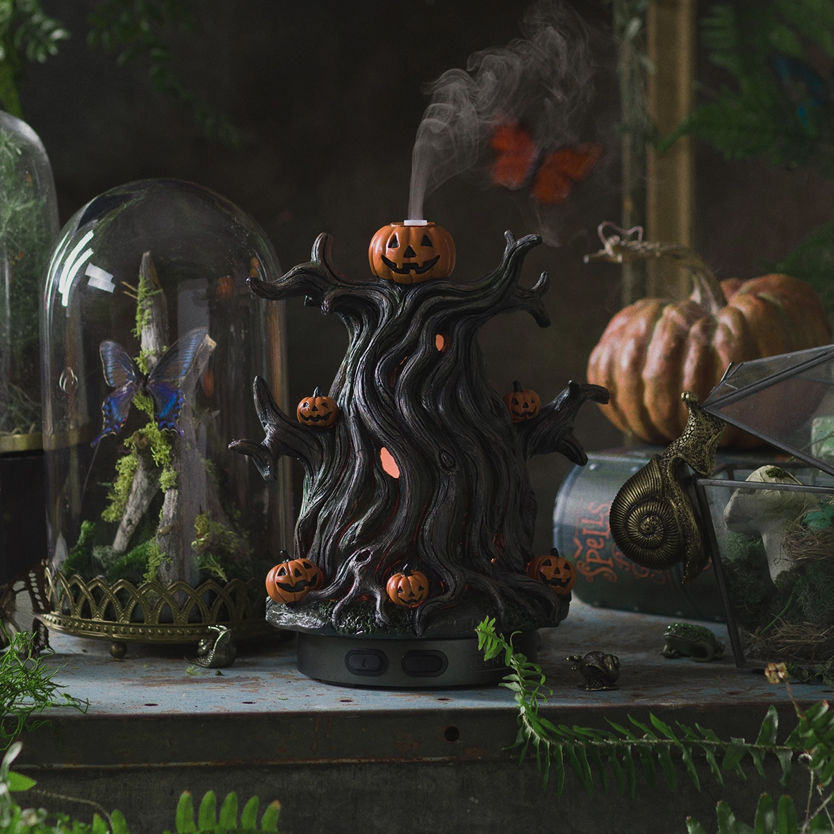 Black gnarled tree-shaped diffuser with jack-o'-lantern pumpkin heads and orange accents, emitting mist, surrounded by Halloween decor including glass domes and dried pumpkins, the Midnight Jack Scentsy Diffuser