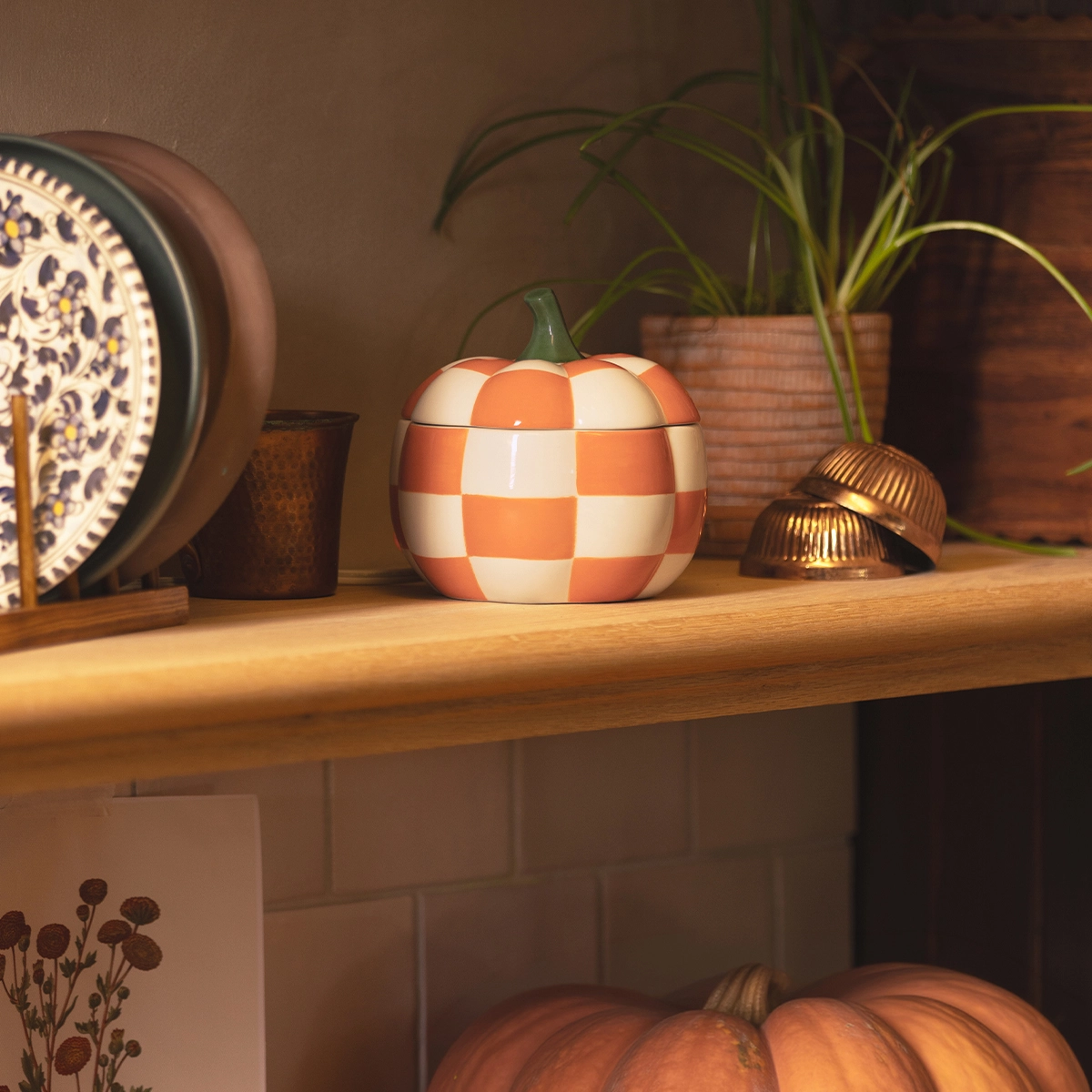 Round ceramic warmer shaped like a pumpkin with orange and cream checkerboard pattern and green stem, displayed on a wooden shelf