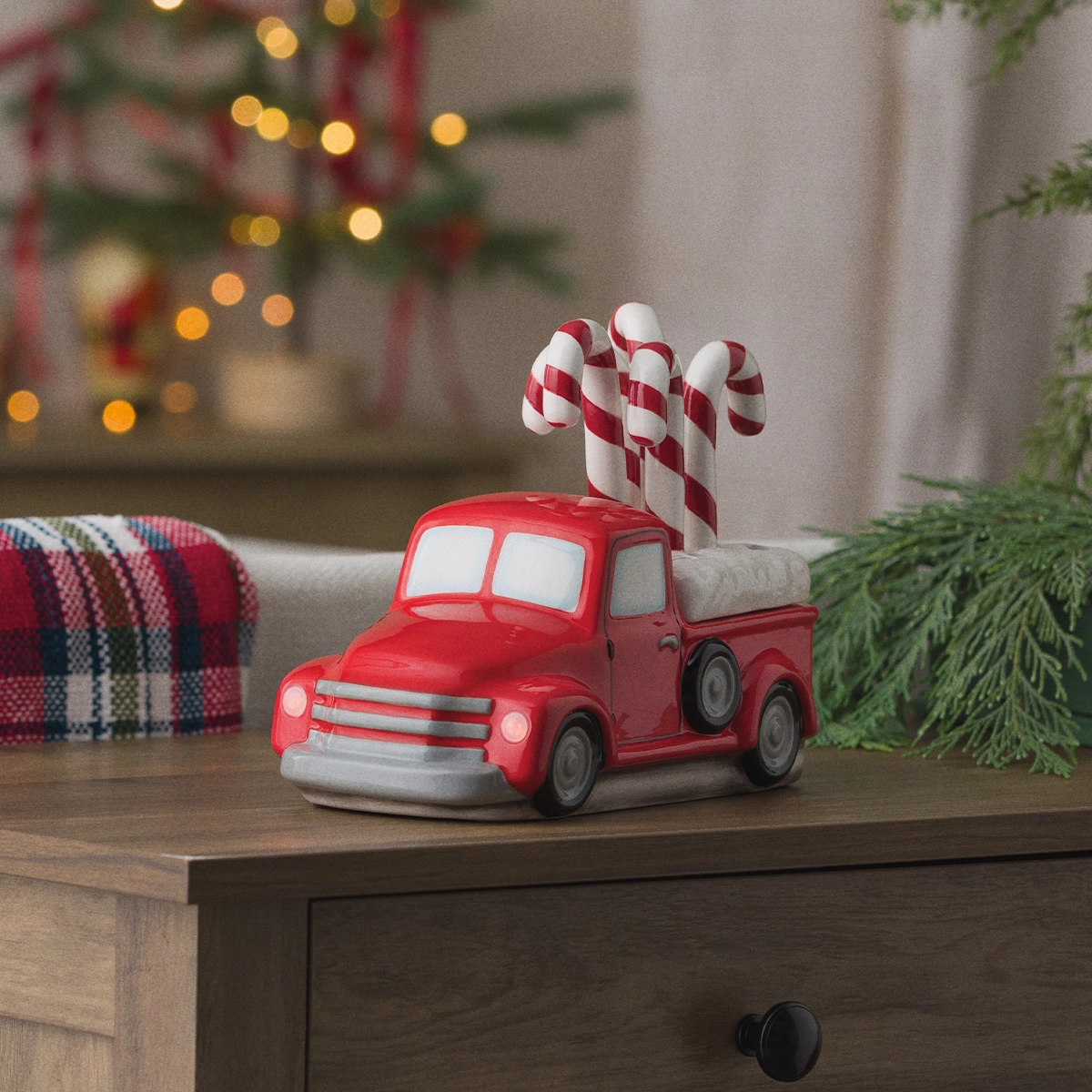 Red retro pickup truck warmer with white-trimmed windows and silver grille, topped with red and white striped candy cane decorations, displayed on a wooden surface surrounded by Christmas greenery and festive lights