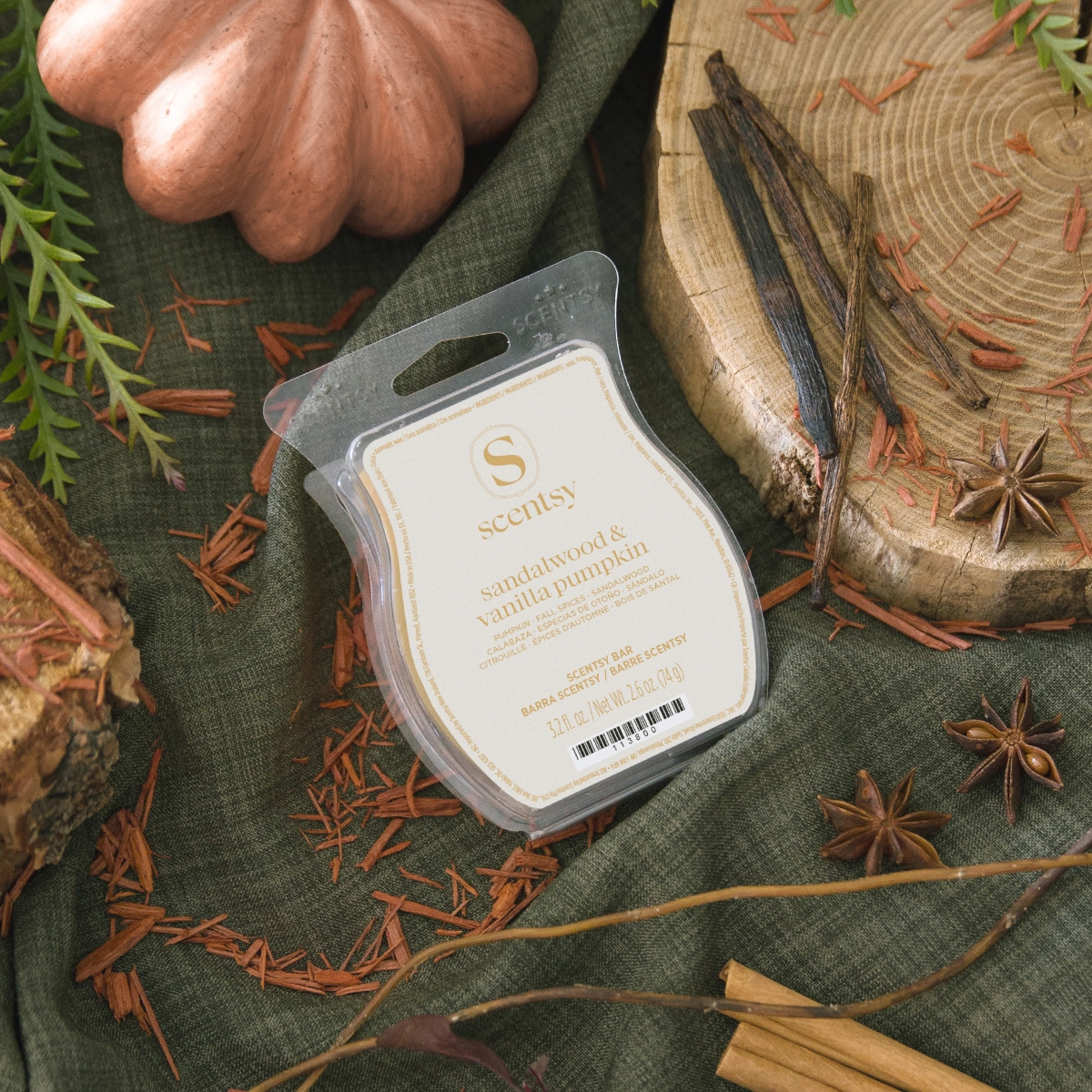 Scentsy Bar wax melt in clear plastic clamshell packaging with cream label and gold text reading "sandalwood & vanilla pumpkin," displayed on a dark green fabric surrounded by dried botanicals, star anise, cinnamon sticks, and a copper pumpkin ornament