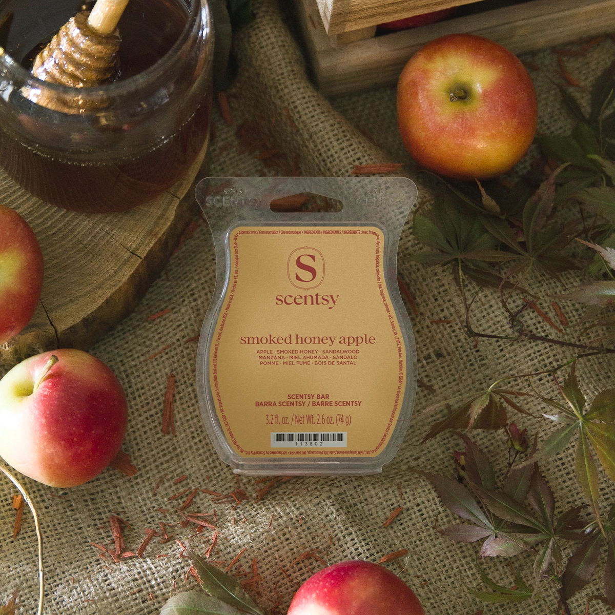 Scentsy Bar in cream packaging with red text labelled "smoked honey apple", surrounded by fresh red apples, dried botanicals, and a glass of dark liquid on textured fabric