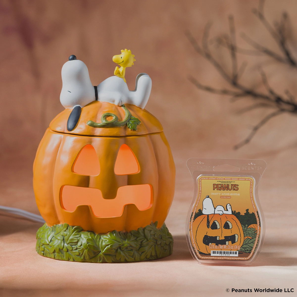 Orange jack-o'-lantern pumpkin warmer with carved face, white dog figurine on top with yellow bird, green leafy base, displayed with Peanuts-themed wax bar