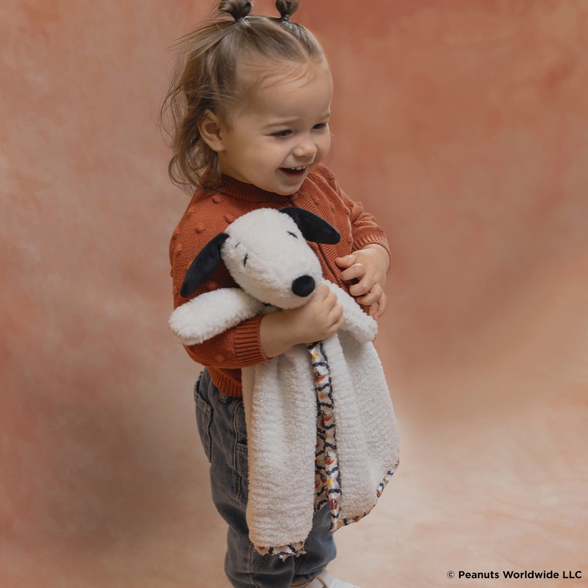 Toddler holding a white plush dog with black ears and nose, wearing an orange knit sweater, against a terracotta background