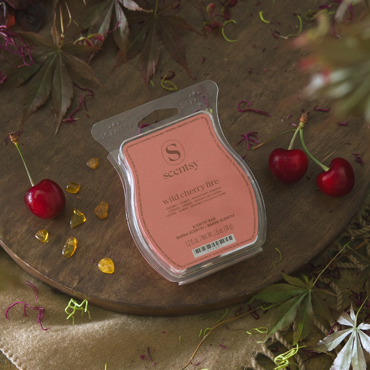 Scentsy Bar in a clamshell case with a coral-pink label showing the Scentsy logo and Wild Cherry Fire scent name, surrounded by fresh cherries, amber gem pieces, and dried flowers on a neutral fabric surface