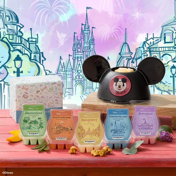 Disney-themed Scentsy product display featuring a black Mickey Mouse ear warmer with red Mickey face emblem, five wax melt packages labelled Adventureland, Frontierland, Main Street, Tomorrowland and Fantasyland in green, orange, yellow, blue and purple, arranged on a pink wooden surface with decorative flowers and foliage, against a pastel illustration of a Disney castle with fireworks and themed buildings.