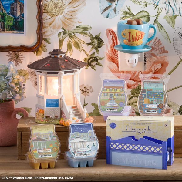 Display of Scentsy Gilmore Girls collection featuring wax melts in decorative packages, a blue teacup warmer, a white lantern-style warmer with black roof, and a blue wooden display box, arranged on wooden shelf against floral wallpaper backdrop.