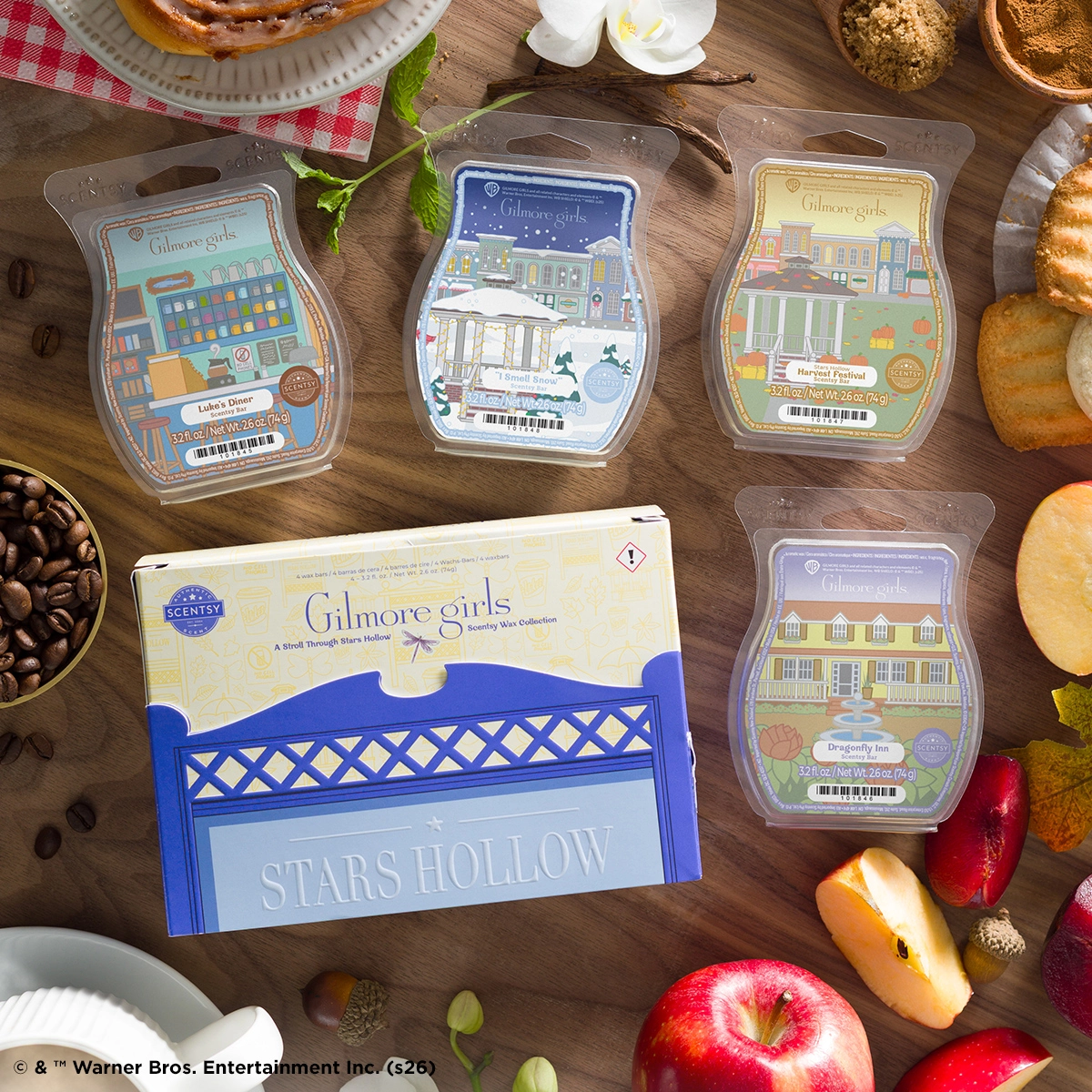 Four Scentsy Wax bars from the Gilmore Girls collection displayed on a wooden surface alongside fresh produce, pastries, and baking ingredients, featuring designs of Stars Hollow locations including Luke's Diner, winter street scene, Harvest Festival, and Dragonfly Inn