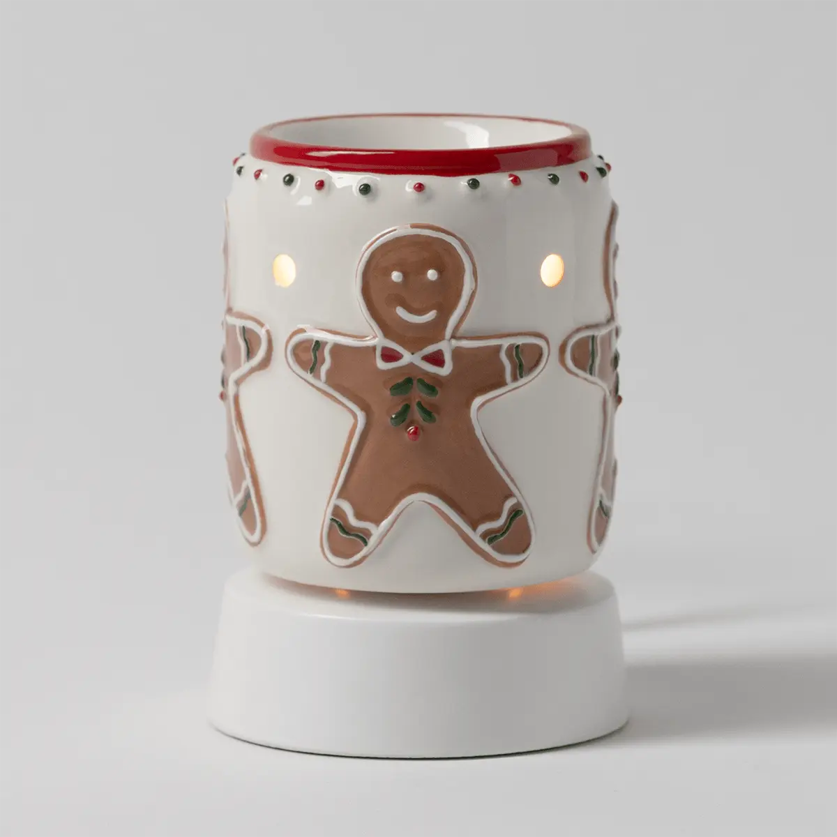 Cream ceramic warmer with gingerbread man design, red trim, and decorative beads, illuminated to show cutout details, the Gingerbread Snap Mini Scentsy Warmer with Tabletop Base