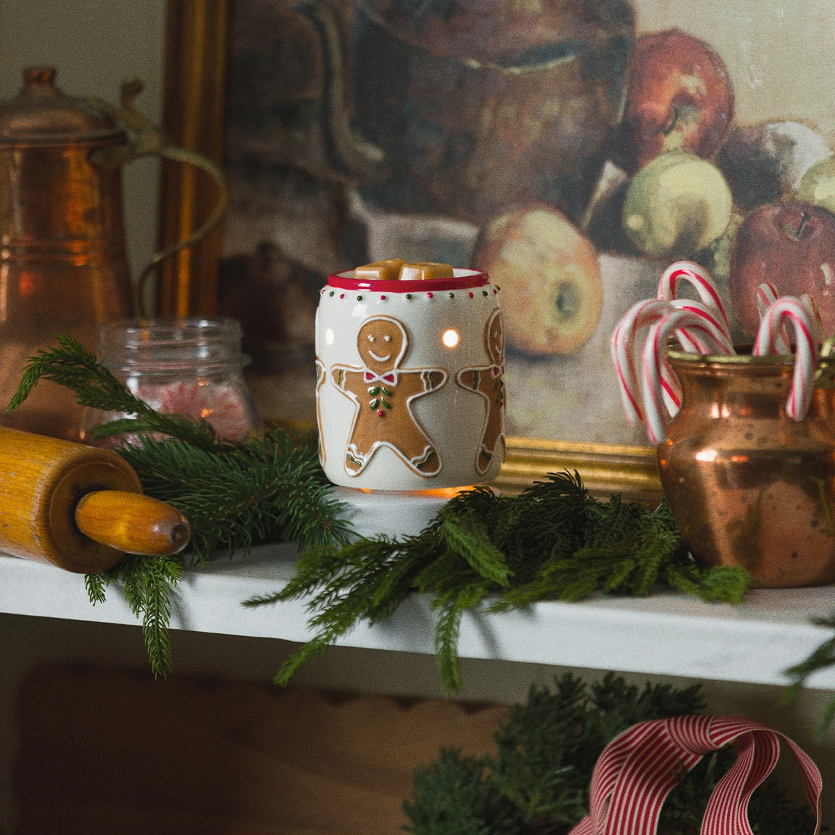 Cylindrical mini warmer with white ceramic base and red rim, decorated with gingerbread man and snowflake designs in brown and gold, surrounded by evergreen sprigs and festive holiday décor