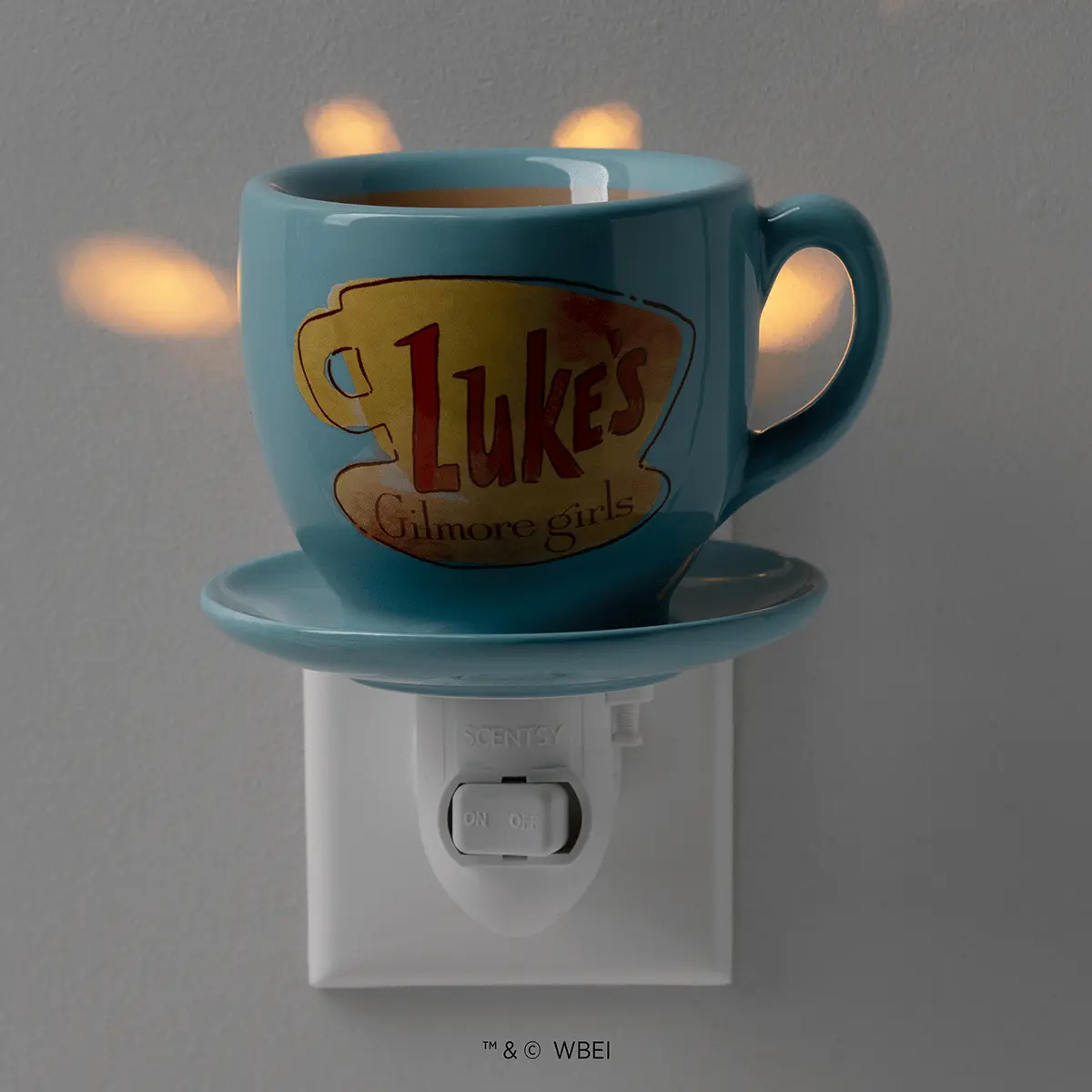 Teal ceramic mug-shaped warmer with "Luke's Gilmore girls" logo, sitting on a matching saucer atop a white wall-plug base with illuminated warmth element, the Luke's Coffee Mug Mini Scentsy Warmer