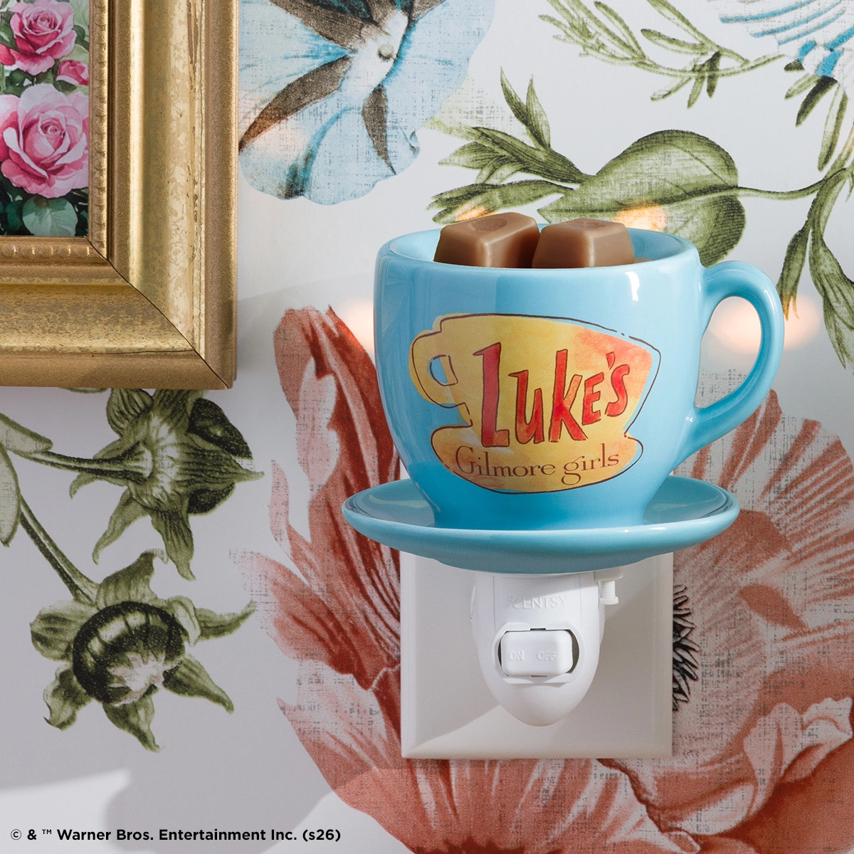 Light blue ceramic mug-shaped warmer with "Luke's Gilmore Girls" logo and matching saucer base, holding two brown wax cubes, mounted on a white wall plug.