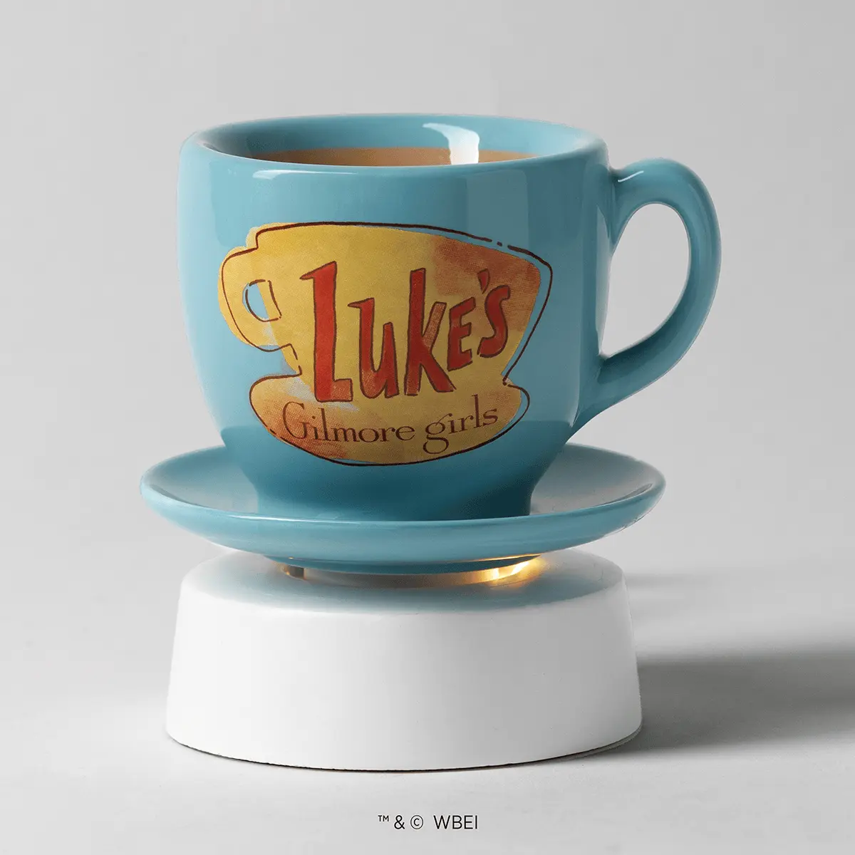 Light blue ceramic mug warmer with a yellow and red "Luke's Coffee Mug" logo and "Gilmore girls" text, seated on a matching saucer with white cylindrical base and gold heating element visible underneath, the Luke’s Coffee Mug Mini Scentsy Warmer with Tabletop Base