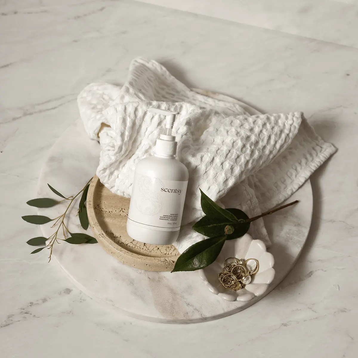 White pump bottle of Pink Coconut Hand Soap on a stone tray with a waffle towel, green leaves and gold rings on marble