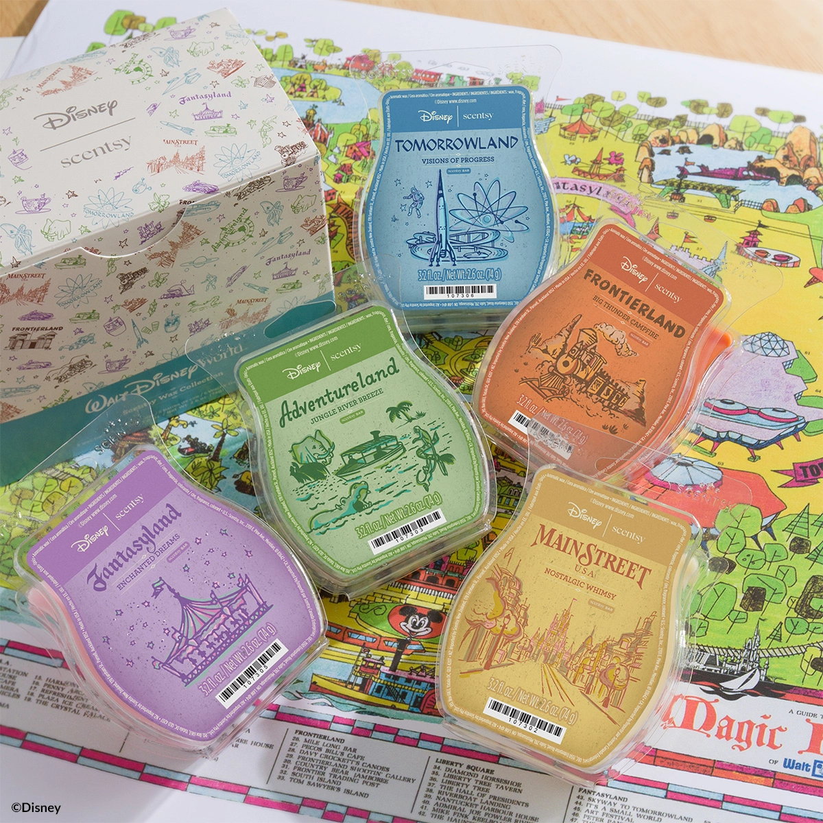 Five Scentsy wax bars in themed clamshell cases featuring Disney Fantasyland, Adventureland, Tomorrowland, Frontierland and Main Street designs, arranged on a colourful Disney park map with a cream Disney-Scentsy branded box in the background