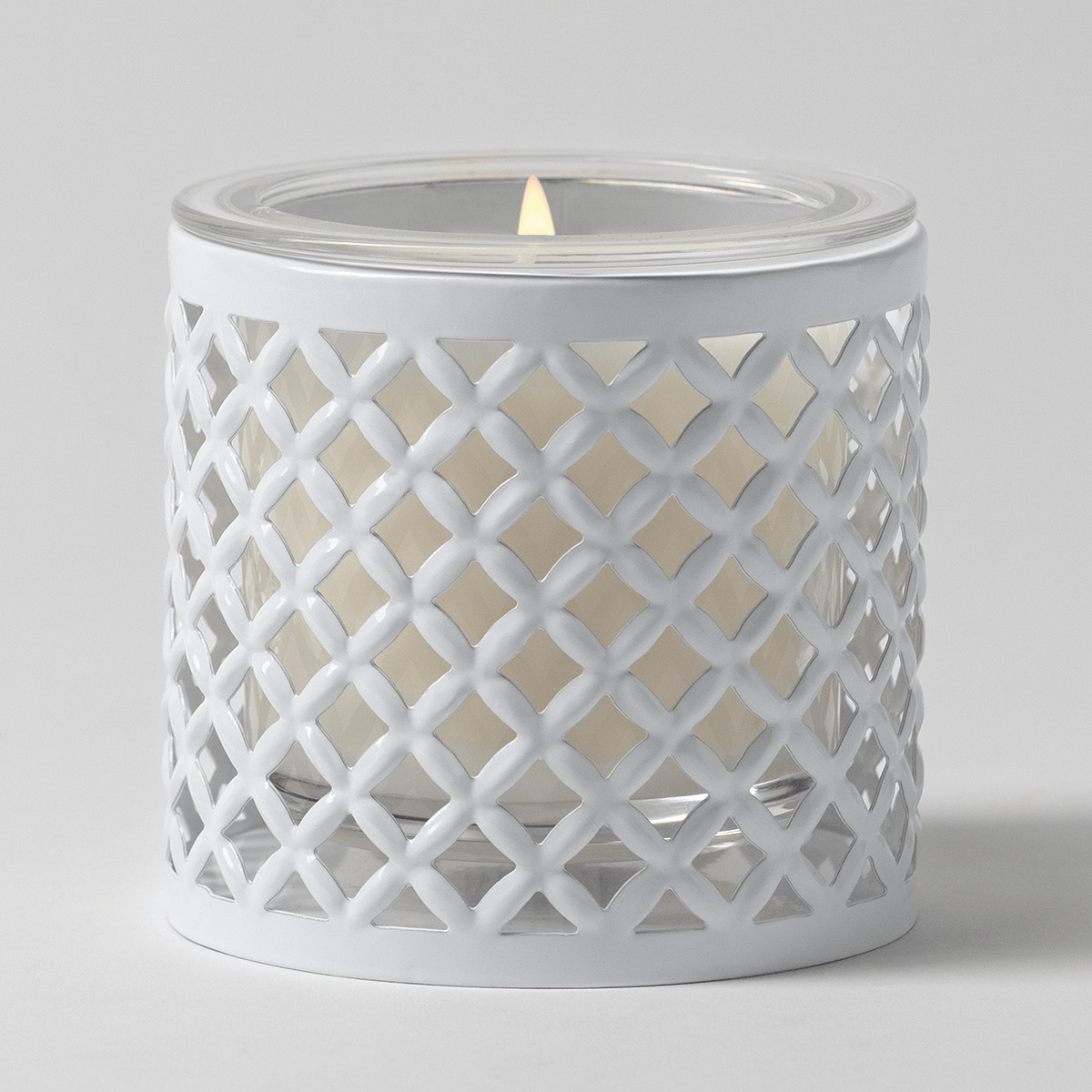 White metal candle holder with an open lattice trellis pattern, holding a clear glass vessel of cream wax with a lit wick