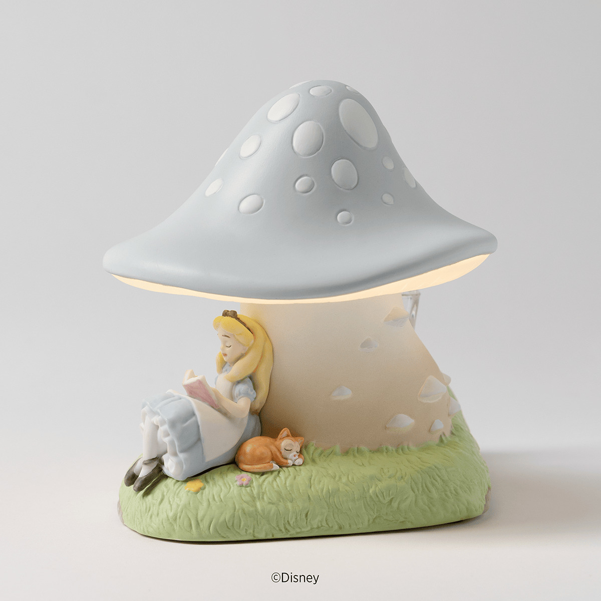 Pale blue spotted ceramic mushroom warmer glowing above Alice reading beside a sleeping kitten on grass, the Disney Alice in Wonderland: Down the Rabbit Hole Scentsy Warmer