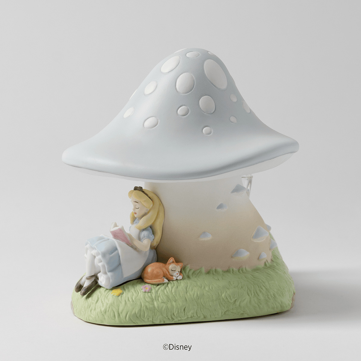 Ceramic warmer shaped like a pale blue spotted mushroom, with Alice reading against its stem and Dinah the cat asleep on grass